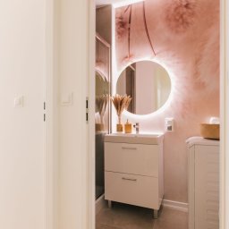 COMFY Biuro Nieruchomości - Nowoczesna łazienka z okrągłym lustrem podświetlanym LED, umywalką z szafką w kolorze złamanej bieli, dekoracyjną tapetą w odcieniach pudrowego różu i beżu oraz suszonymi trawami w wazonie.