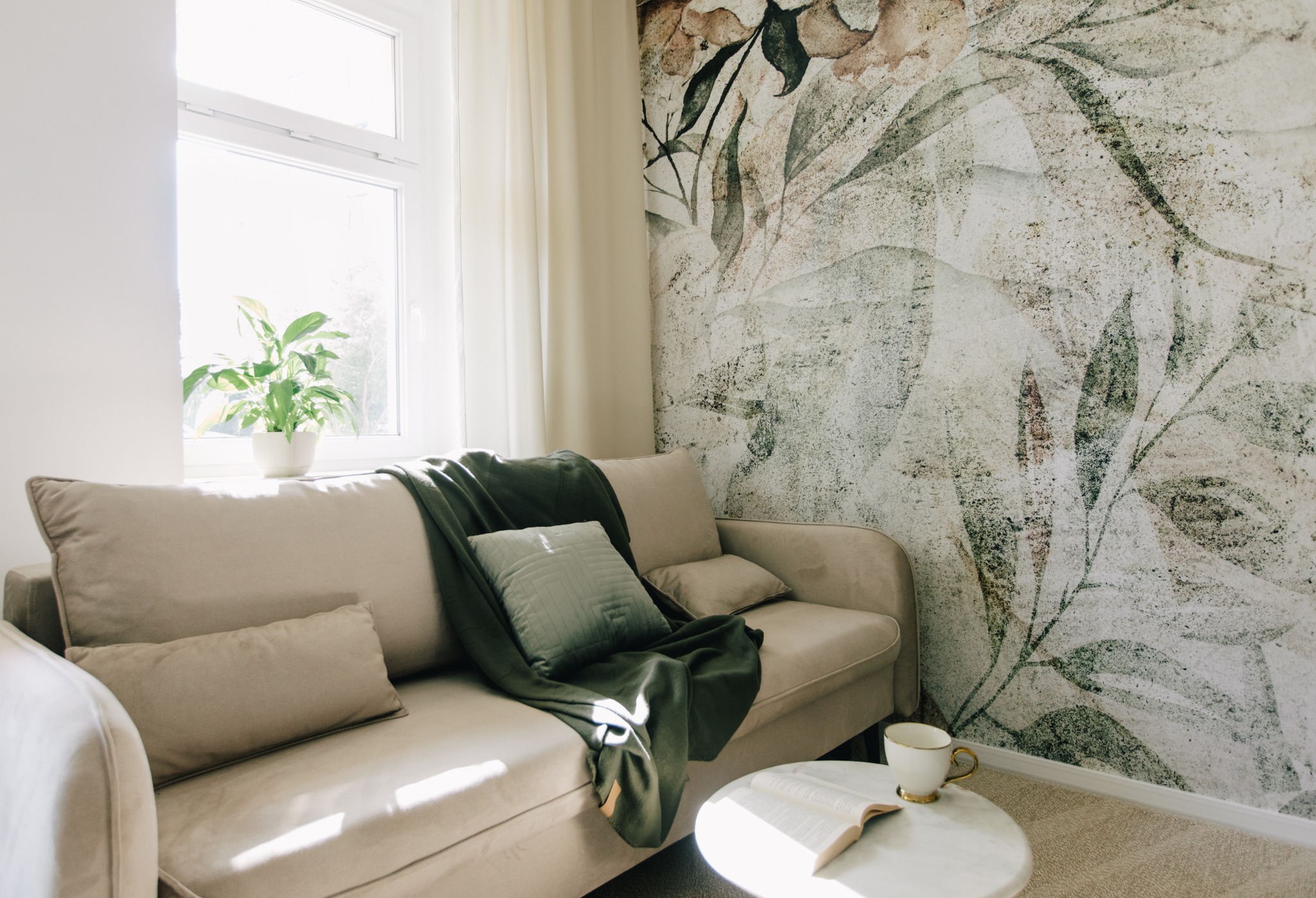 Jasny salon z beżową sofą, zielonym pledem i poduszkami, stolikiem kawowym z książką i kubkiem, oraz fototapetą w motywy roślinne.