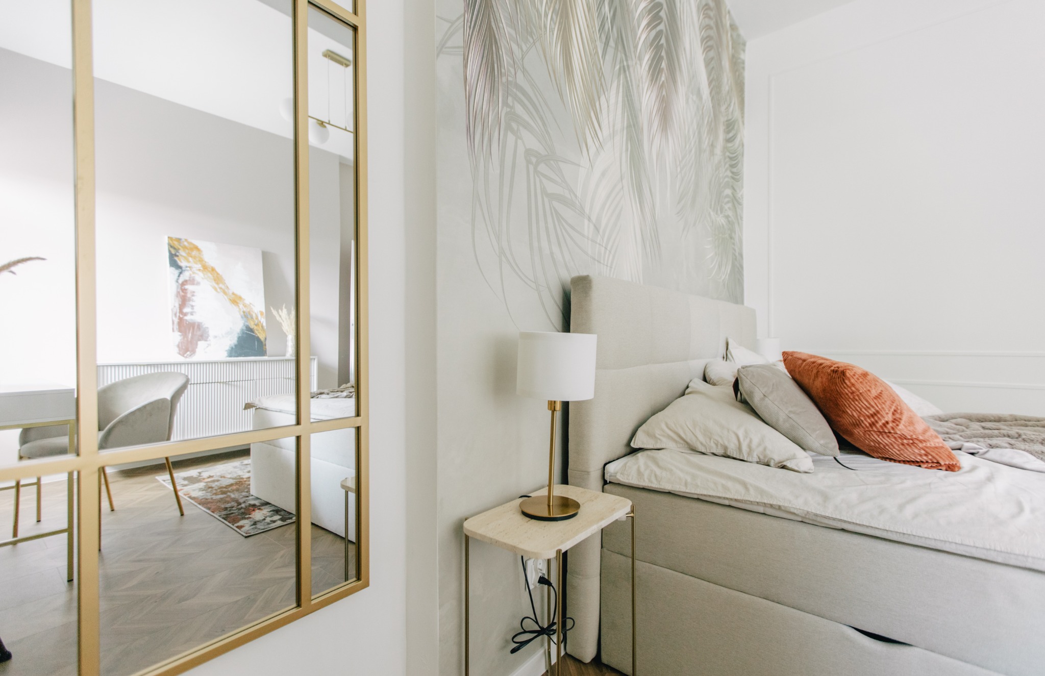 Jasna sypialnia z łóżkiem kontynentalnym, stolikiem nocnym z lampką i dużym lustrem w złotej ramie, w którym odbija się część pokoju z biurkiem i krzesłem.