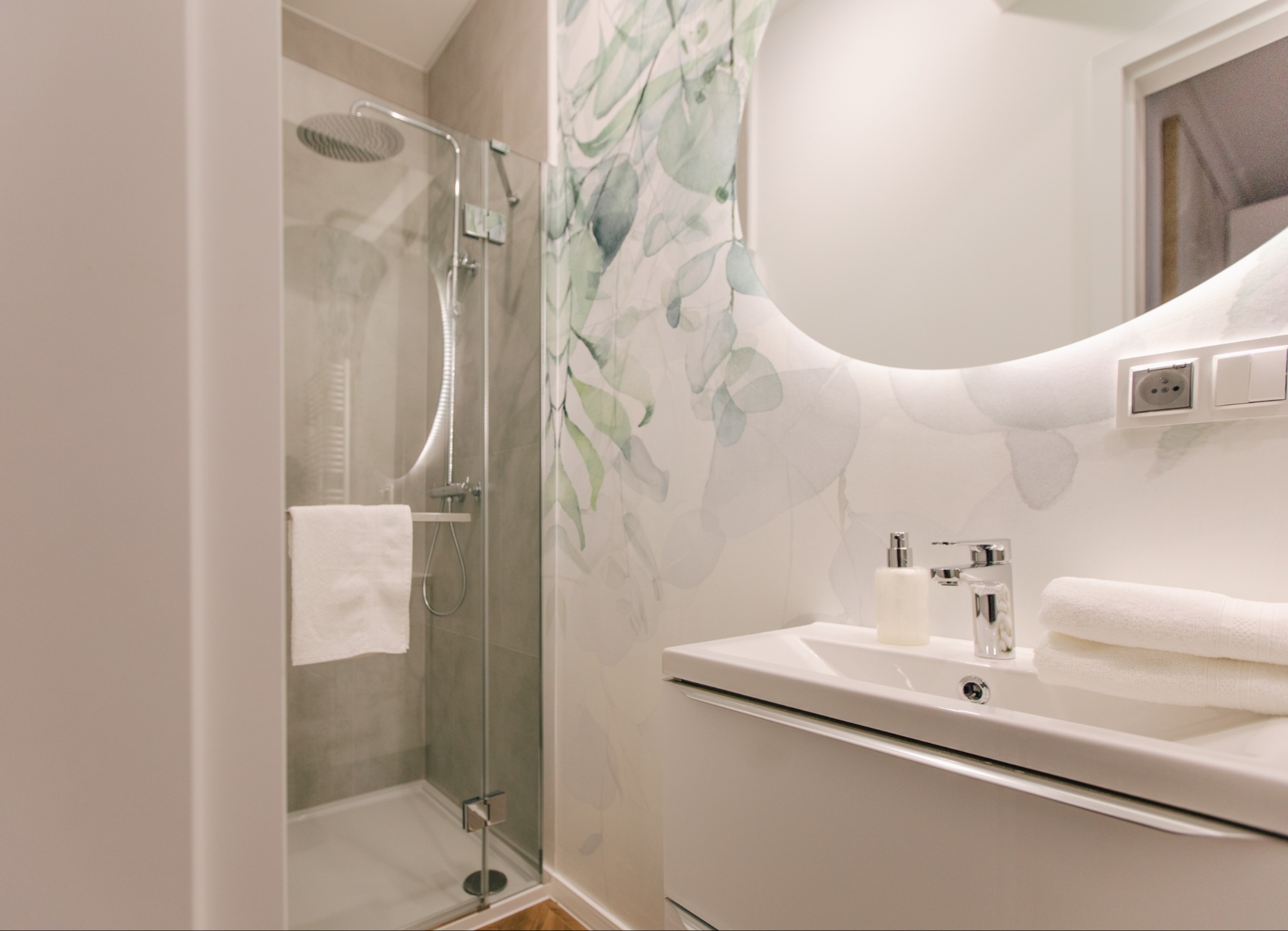 Nowoczesna łazienka z kabiną prysznicową, okrągłym lustrem z podświetleniem LED, umywalką z szafką i akcentem roślinnym na ścianie.