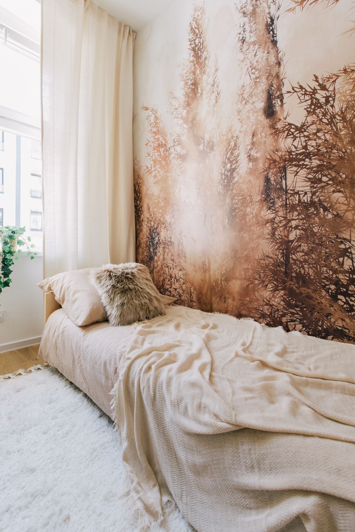 Jasna sypialnia z łóżkiem, puszystą poduszką i fototapetą z motywem traw na ścianie, obok okna roślina doniczkowa.