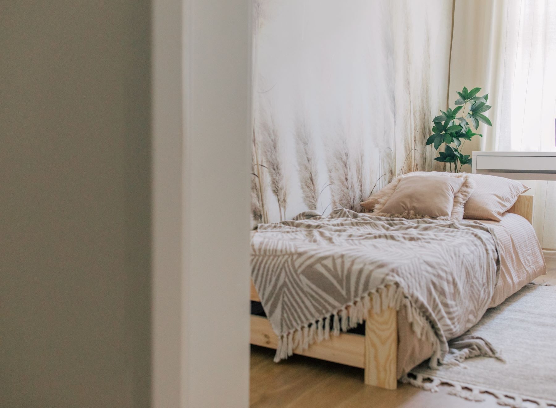 Jasna sypialnia z drewnianym łóżkiem, beżową pościelą i poduszkami, tapeta z motywem traw, zielona roślina na białej komodzie, widziana zza framugi drzwi.