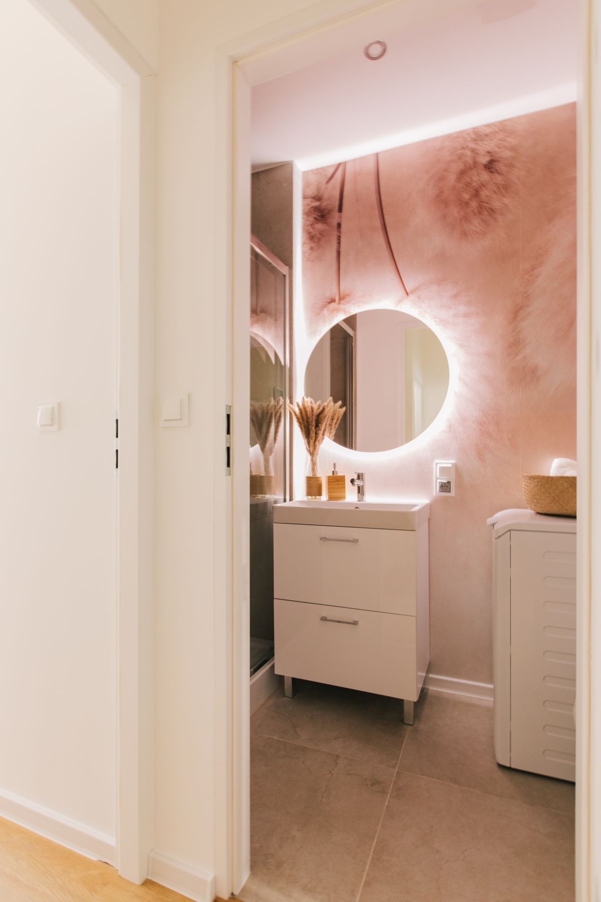 Nowoczesna łazienka z okrągłym lustrem podświetlanym LED, umywalką z szafką w kolorze złamanej bieli, dekoracyjną tapetą w odcieniach pudrowego różu i beżu oraz suszonymi trawami w wazonie.