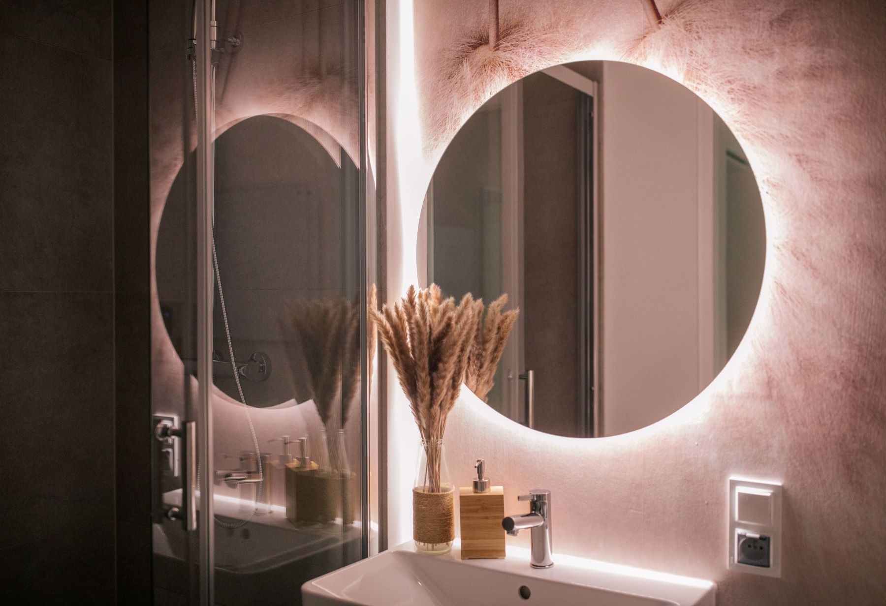 Nowoczesna łazienka z okrągłym lustrem podświetlanym LED, umywalką, baterią, dozownikiem mydła z bambusa i dekoracyjnym bukietem suszonych traw w szklanym wazonie owiniętym sznurkiem.