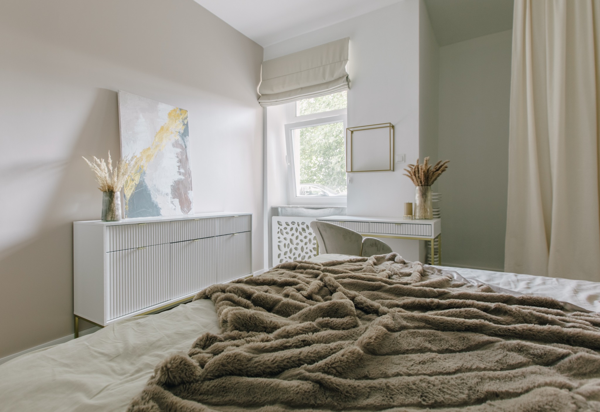 Jasna sypialnia z łóżkiem przykrytym puszystym, brązowym kocem, biała komoda z abstrakcyjnym obrazem i wazonem z suszem, toaletka z krzesłem i dekoracjami, okno z roletą rzymską.