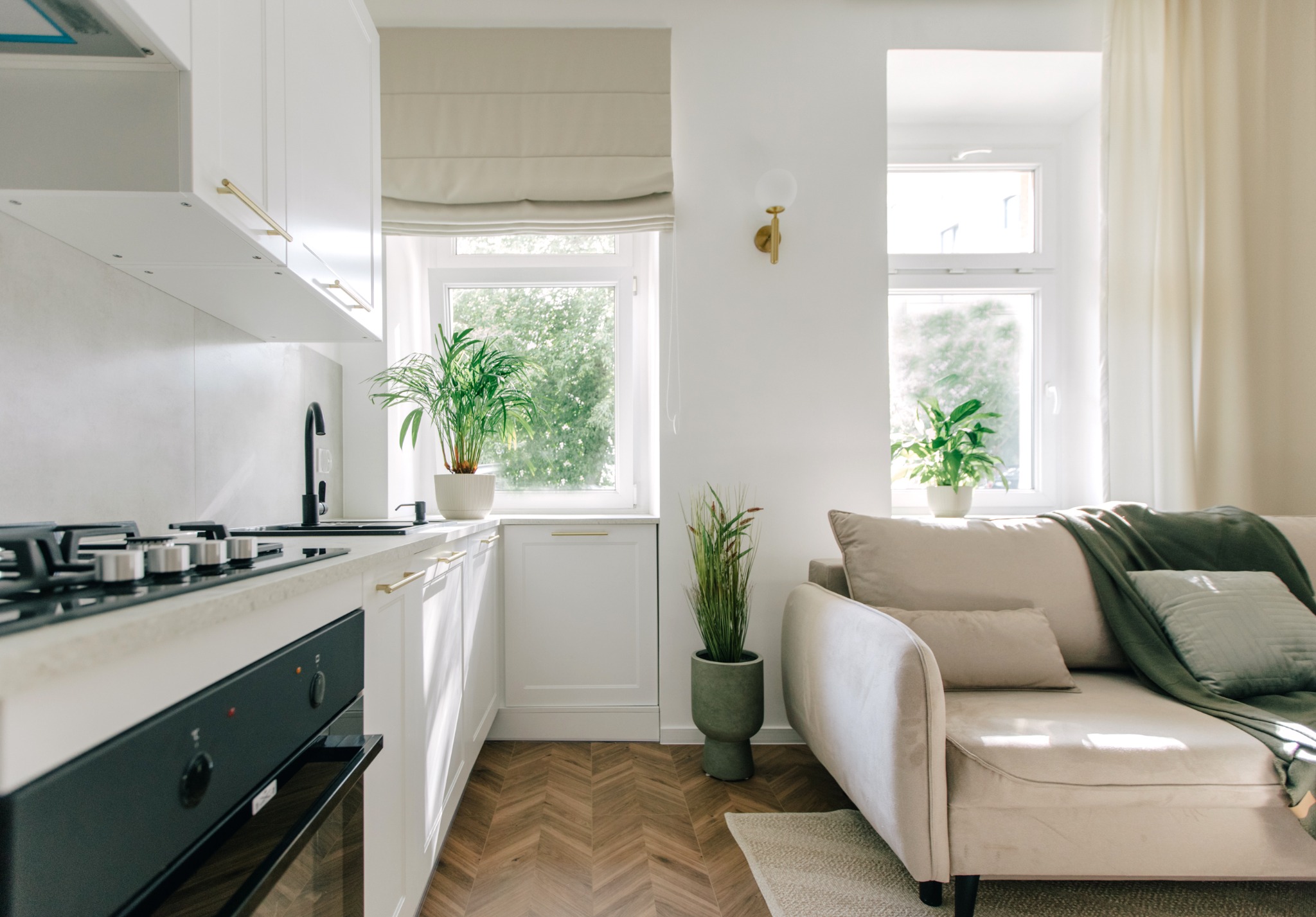 Jasne wnętrze mieszkania z aneksem kuchennym i wypoczynkowym, drewniana podłoga w jodełkę, białe meble kuchenne z złotymi uchwytami, czarna armatura, beżowa sofa z zielonym pledem.
