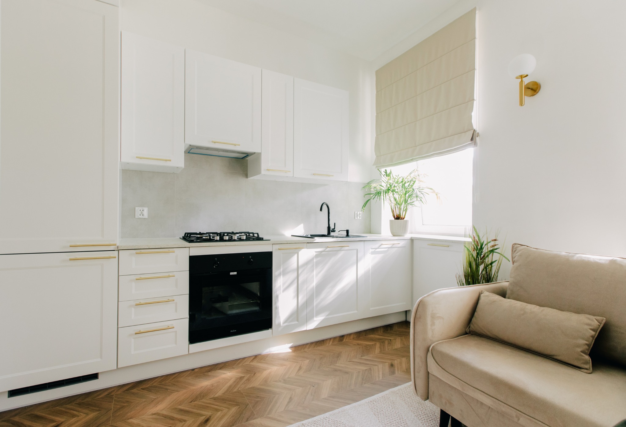 Jasna kuchnia z białymi szafkami, złotymi uchwytami, czarną armaturą i parkietem w jodełkę, widoczny fragment beżowej sofy