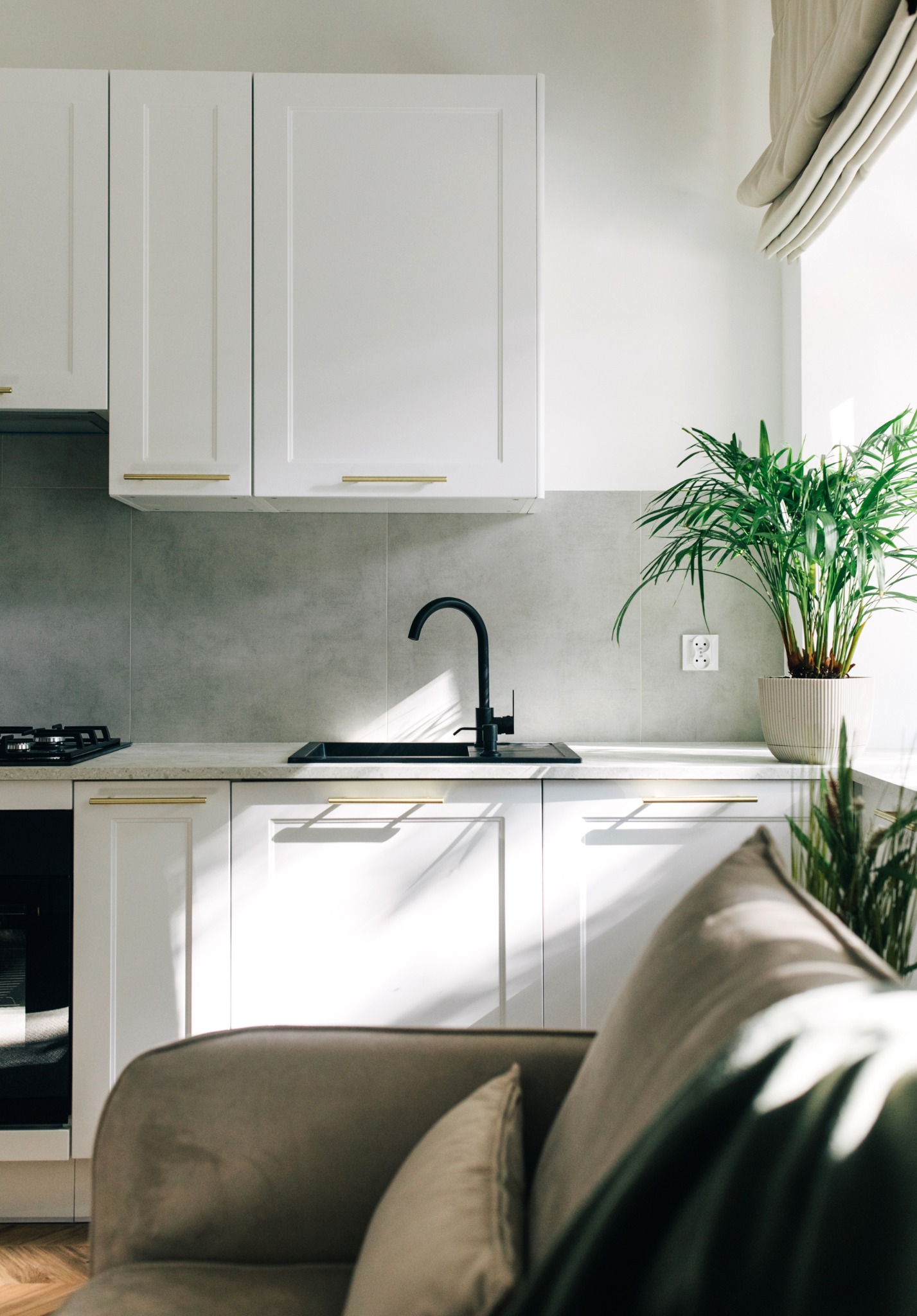 Jasna kuchnia z białymi szafkami, złotymi uchwytami i czarną armaturą, uzupełniona o zieloną roślinę doniczkową na blacie, z fragmentem sofy w odcieniach beżu na pierwszym planie.