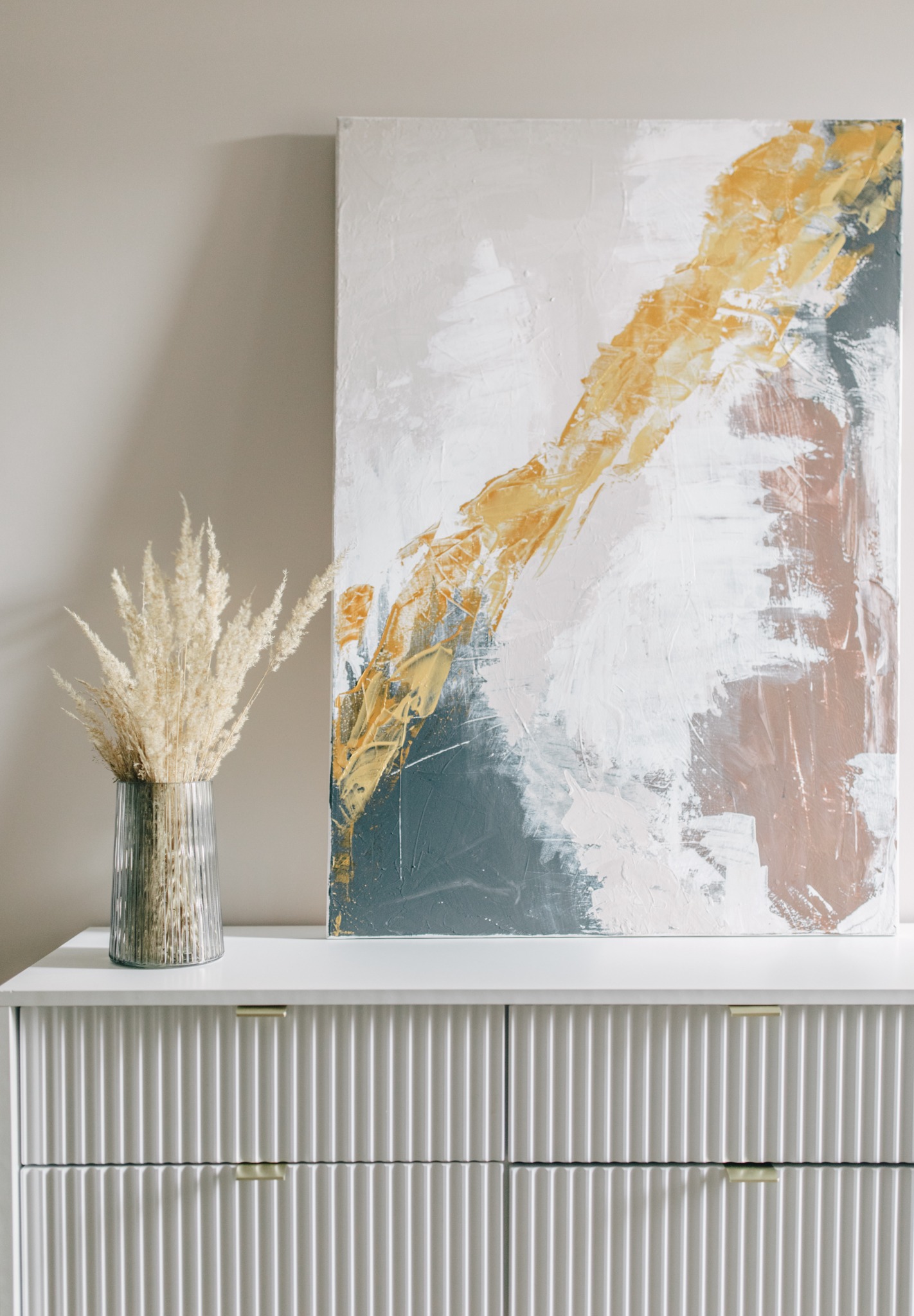 Abstrakcyjny obraz z przewagą bieli, złota i szarości, oparty o białą komodę z ryflowanymi frontami szuflad; obok wazon z suszonymi trawami w odcieniach beżu.