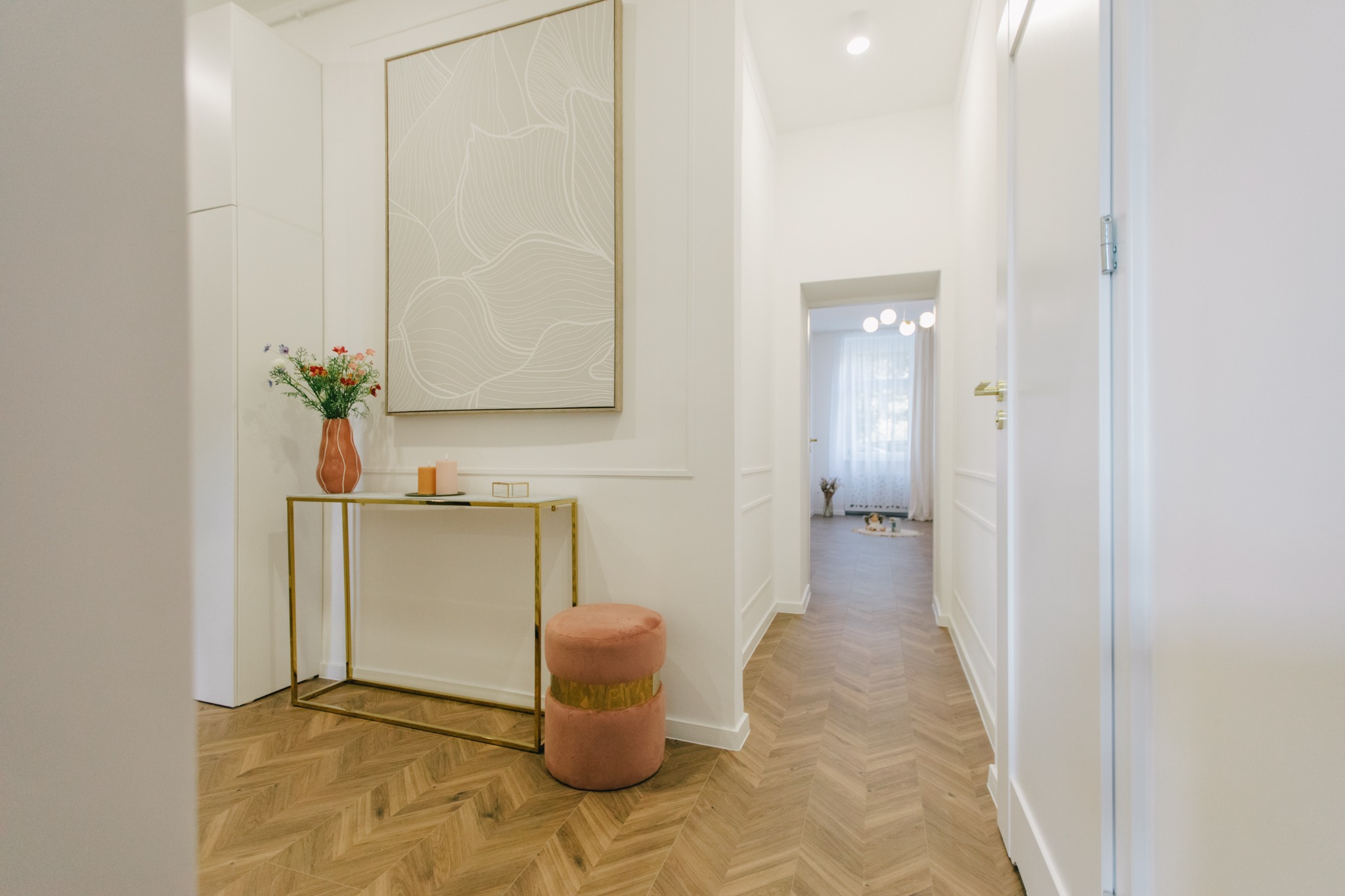 Jasny przedpokój z parkietem w jodełkę, konsolą z bukietem kwiatów i abstrakcyjnym obrazem, uzupełniony pufem w kolorze pudrowego różu i widokiem na dalszą część mieszkania.