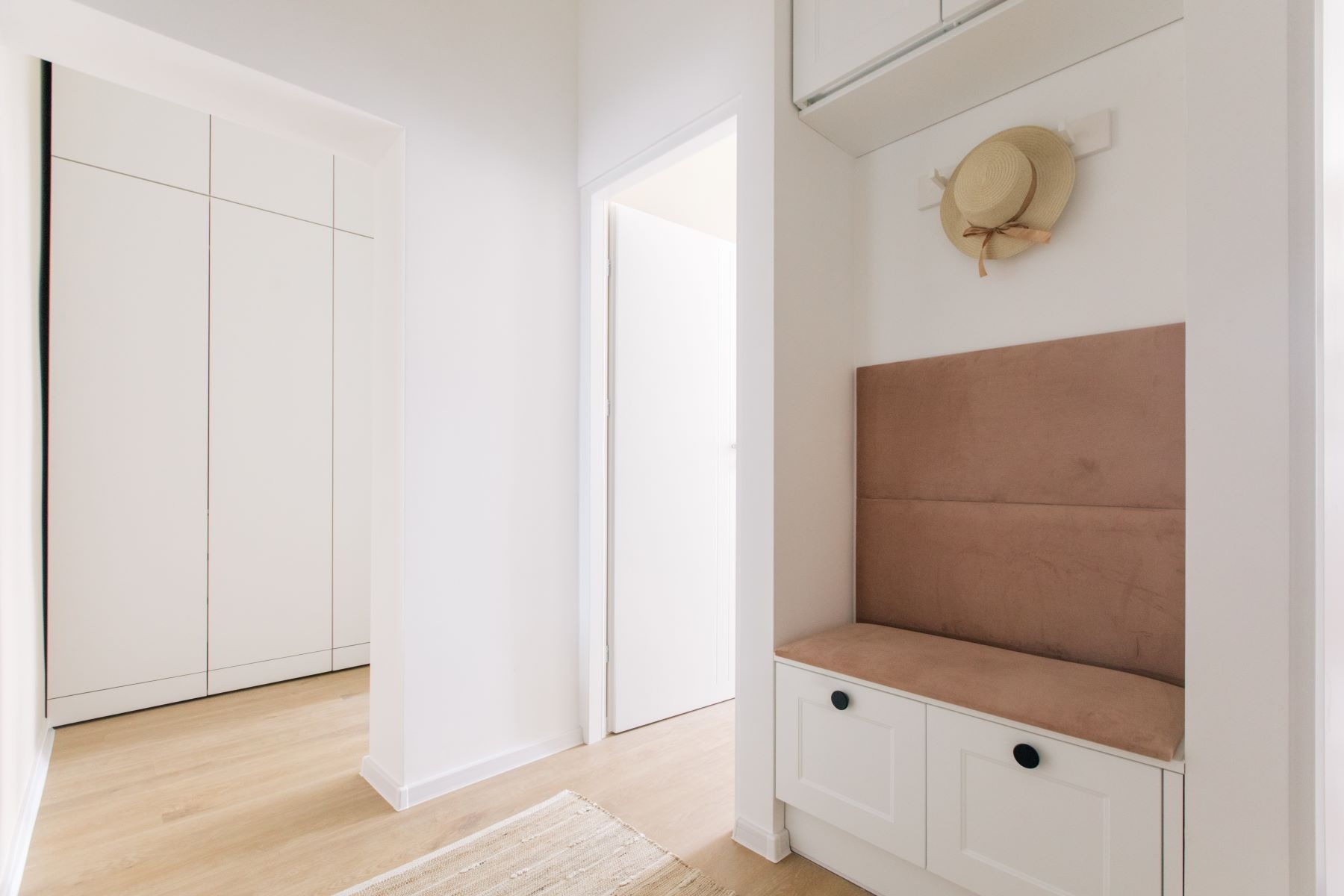 Jasny przedpokój z białą zabudową szaf, siedziskiem z brązowym obiciem, słomkowym kapeluszem na wieszaku i jasnym dywanikiem na podłodze.