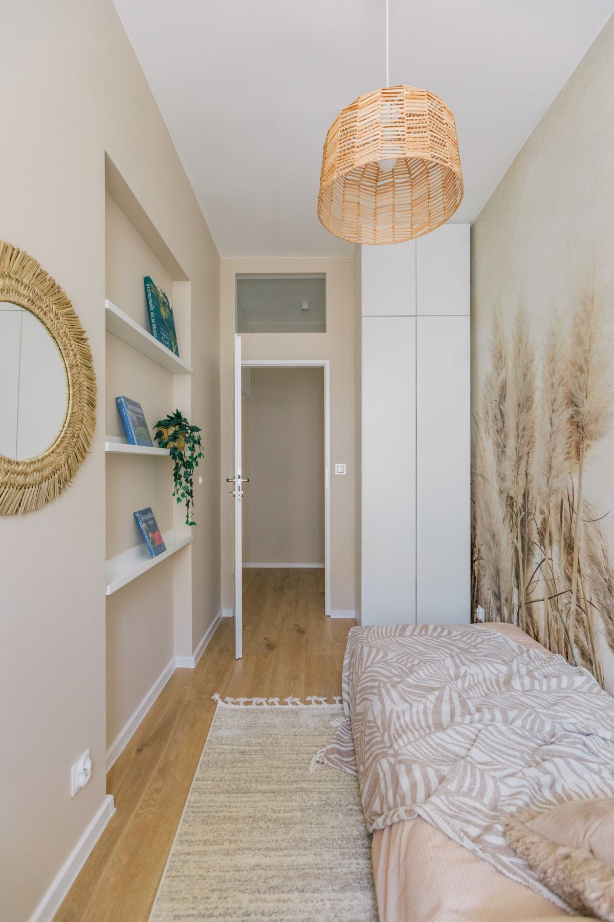 Beżowa sypialnia z łóżkiem, tapetą w trawy pampasowe, wiklinowym lustrem i lampą oraz otwartymi półkami z książkami i rośliną.