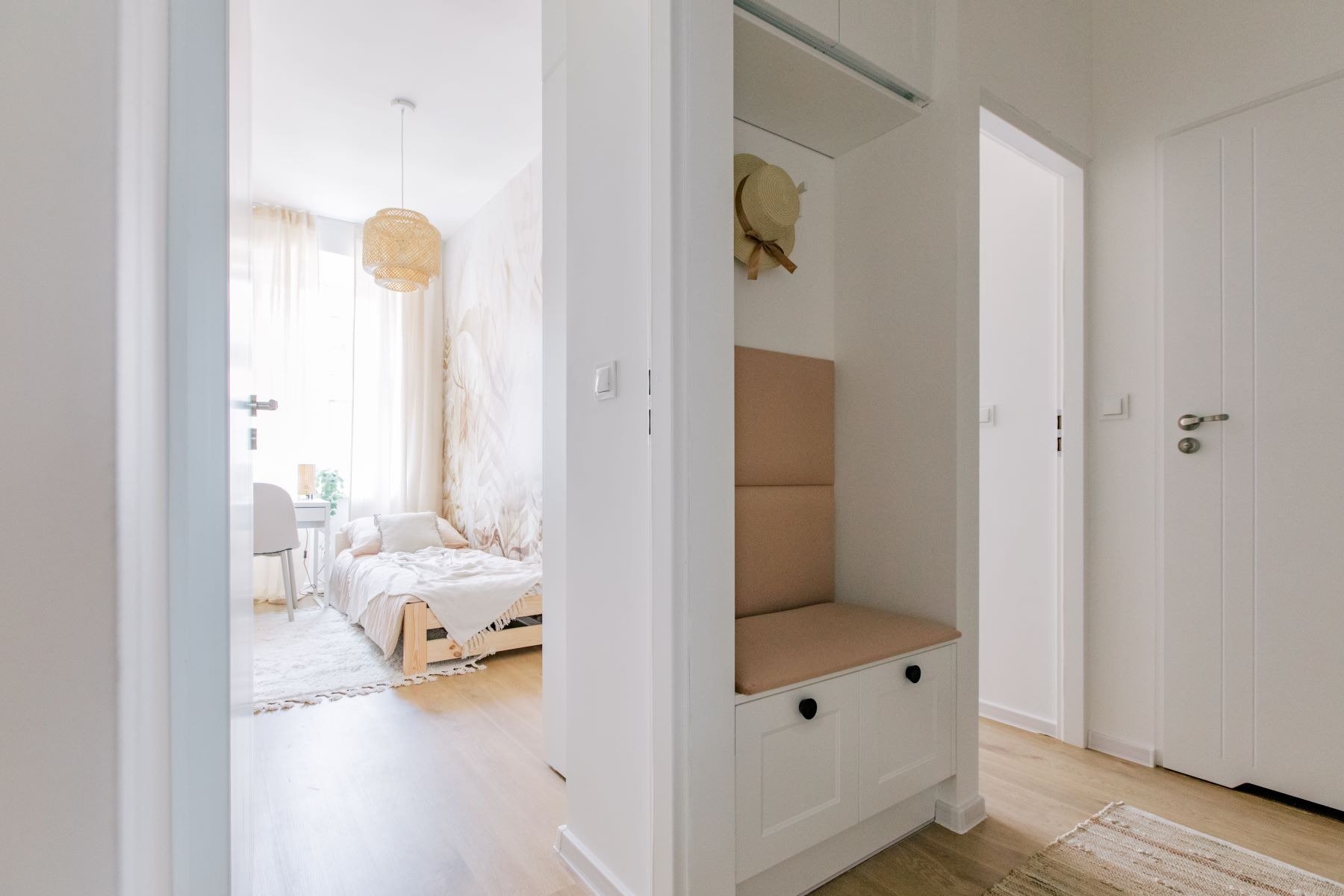 Jasne wnętrze: widok na pokój z łóżkiem i biurkiem przez drzwi, obok wnęka z siedziskiem i szafkami, słomkowy kapelusz na wieszaku, dywanik na podłodze.