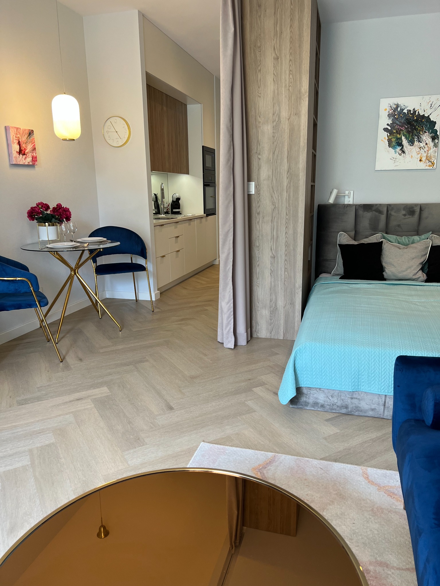 Wnętrze kawalerki z aneksem kuchennym, stołem z kwiatami i łóżkiem za drewnianą ścianką działową, w neutralnej kolorystyce z akcentami złota i granatu.