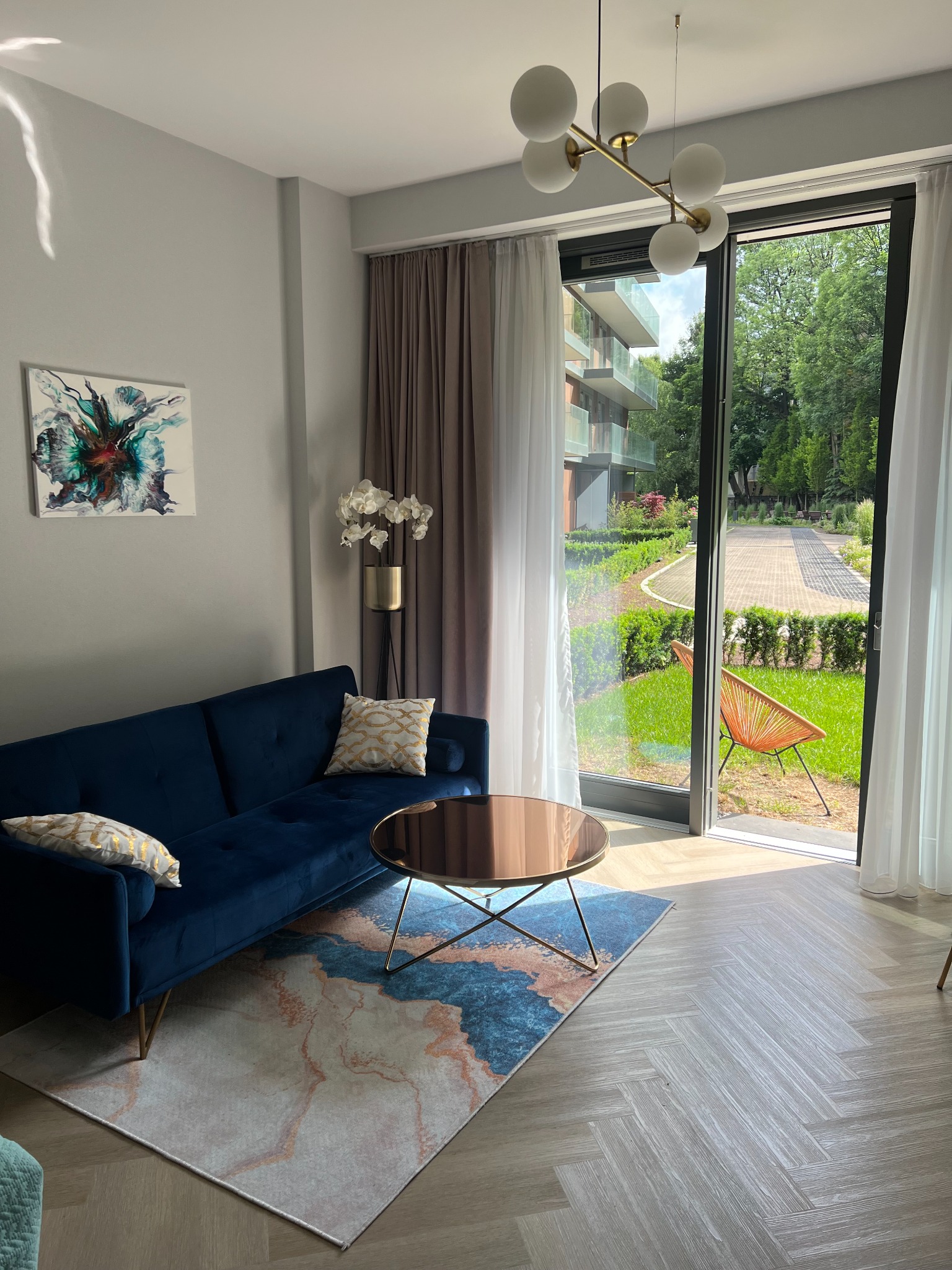 Nowoczesny salon z granatową sofą, okrągłym stolikiem kawowym i widokiem na zadbany ogród z krzesłem przez przesuwne szklane drzwi.