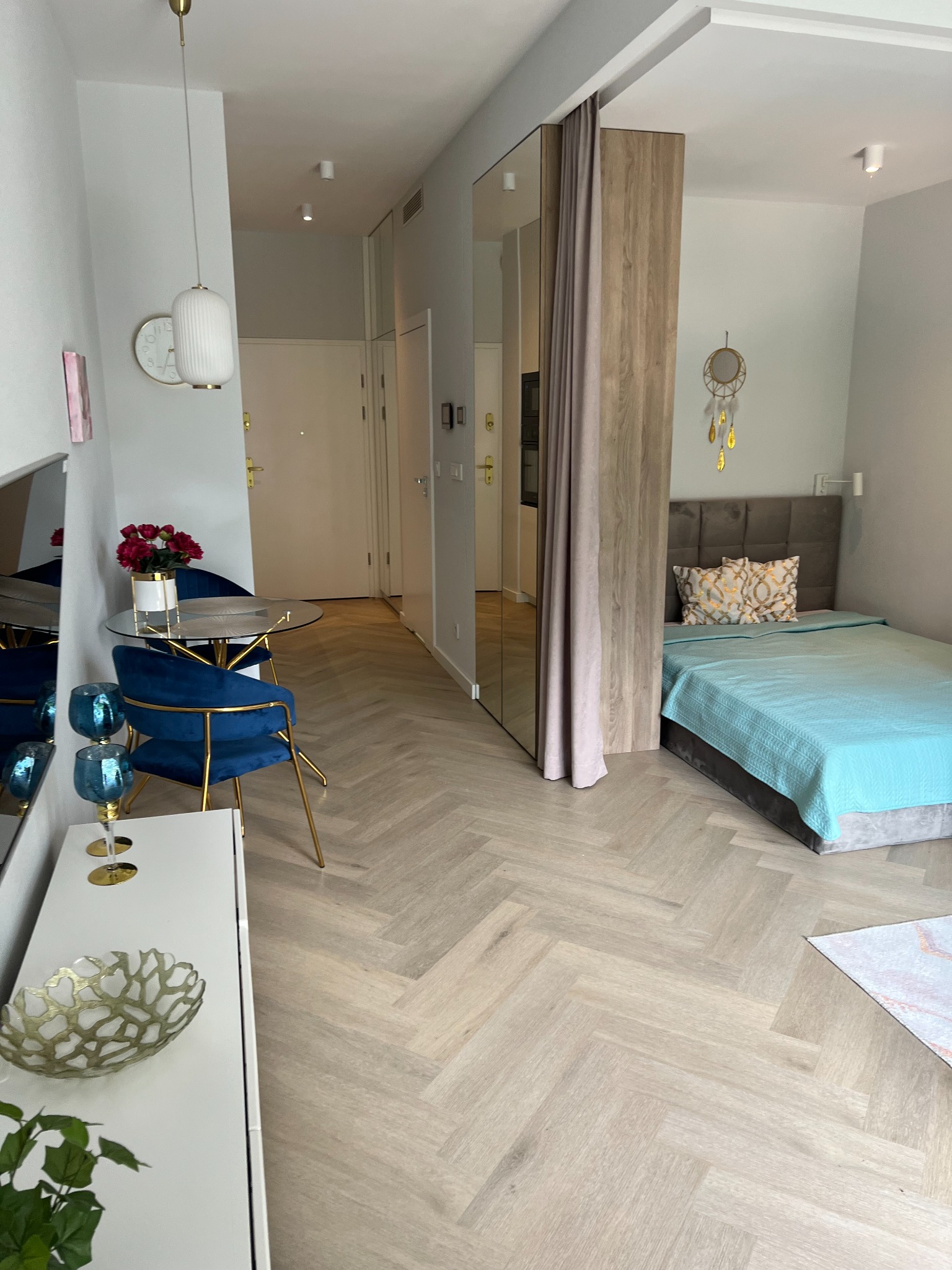 Nowoczesne studio: sypialnia z łóżkiem z turkusową narzutą oddzielona od części dziennej z okrągłym szklanym stołem i granatowymi krzesłami, podłoga z jasnego parkietu ułożonego w jodełkę.