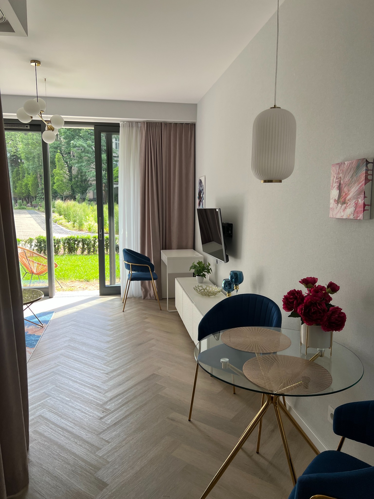 Jasne wnętrze mieszkania z parkietem w jodełkę, szklanym stołem z złotymi nogami, granatowymi krzesłami, telewizorem na ścianie i widokiem na ogród przez przeszklone drzwi balkonowe.