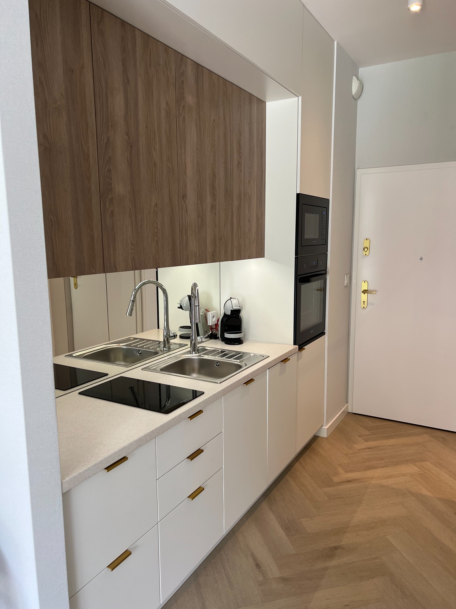 Nowoczesna kuchnia z drewnianymi szafkami wiszącymi, białymi szafkami dolnymi z poziomymi złotymi uchwytami, podwójnym zlewem ze stali nierdzewnej, czarną płytą indukcyjną i parkietem w jodełkę...