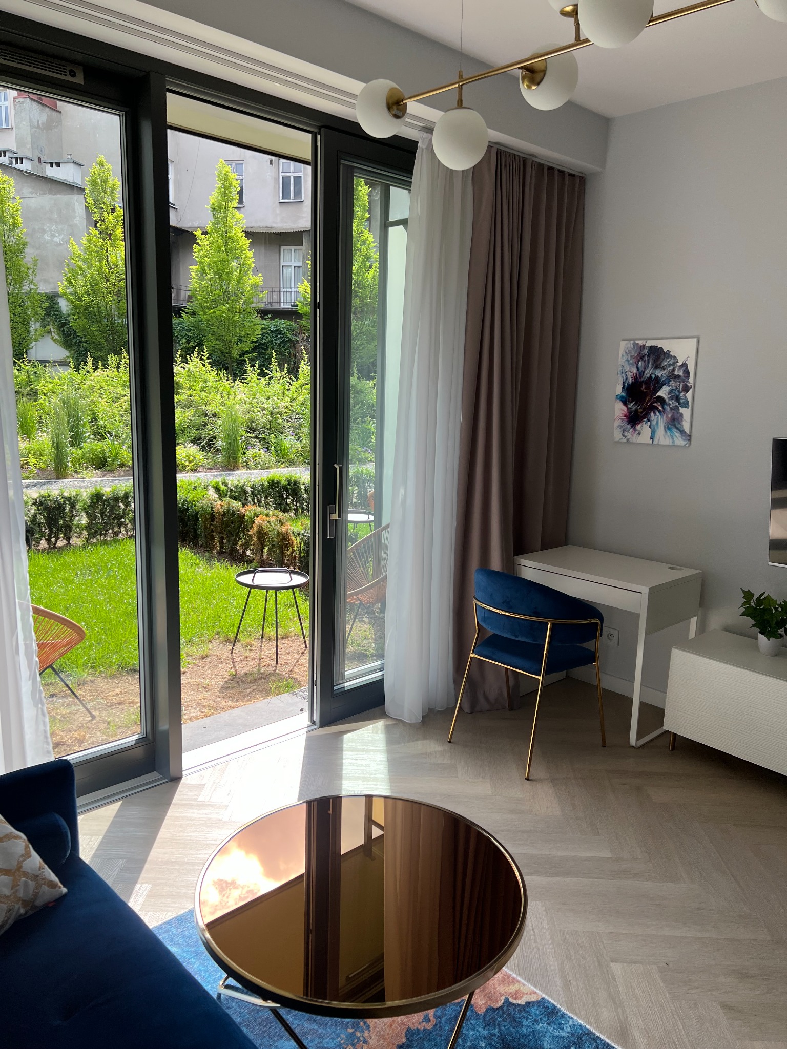 Nowoczesne wnętrze mieszkania z widokiem na zielony ogród przez duże okno balkonowe, okrągły stolik kawowy z lustrzanym blatem odbija światło.