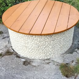 PavSorBud - Okrągły stolik z drewnianym blatem i podstawą wykończoną białymi kamykami na betonowym podłożu, otoczony zielenią.