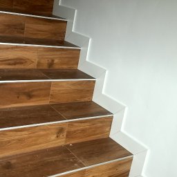 PavSorBud - Schody wyłożone brązowymi płytkami imitującymi drewno, z białymi listwami przypodłogowymi, widok z dołu.