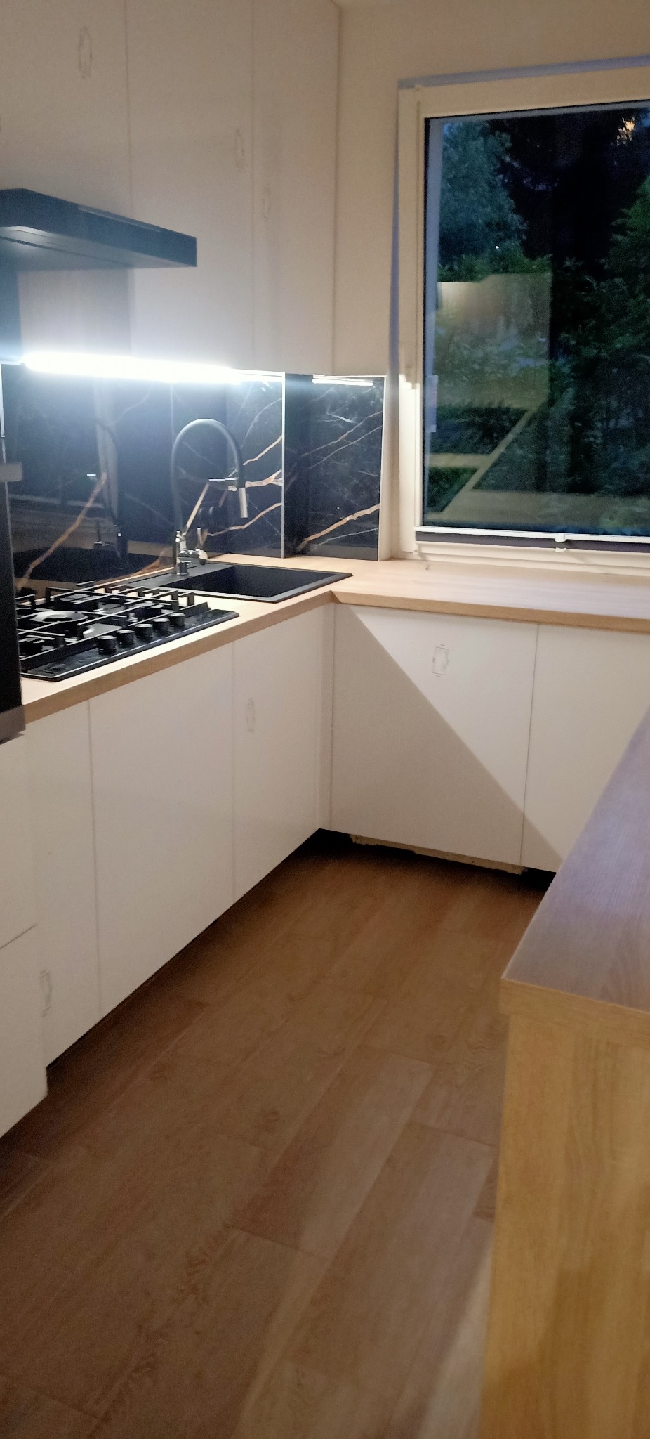 Nowoczesna kuchnia z białymi szafkami, drewnianym blatem i czarnym panelem ściennym imitującym marmur, oświetlonym od góry.