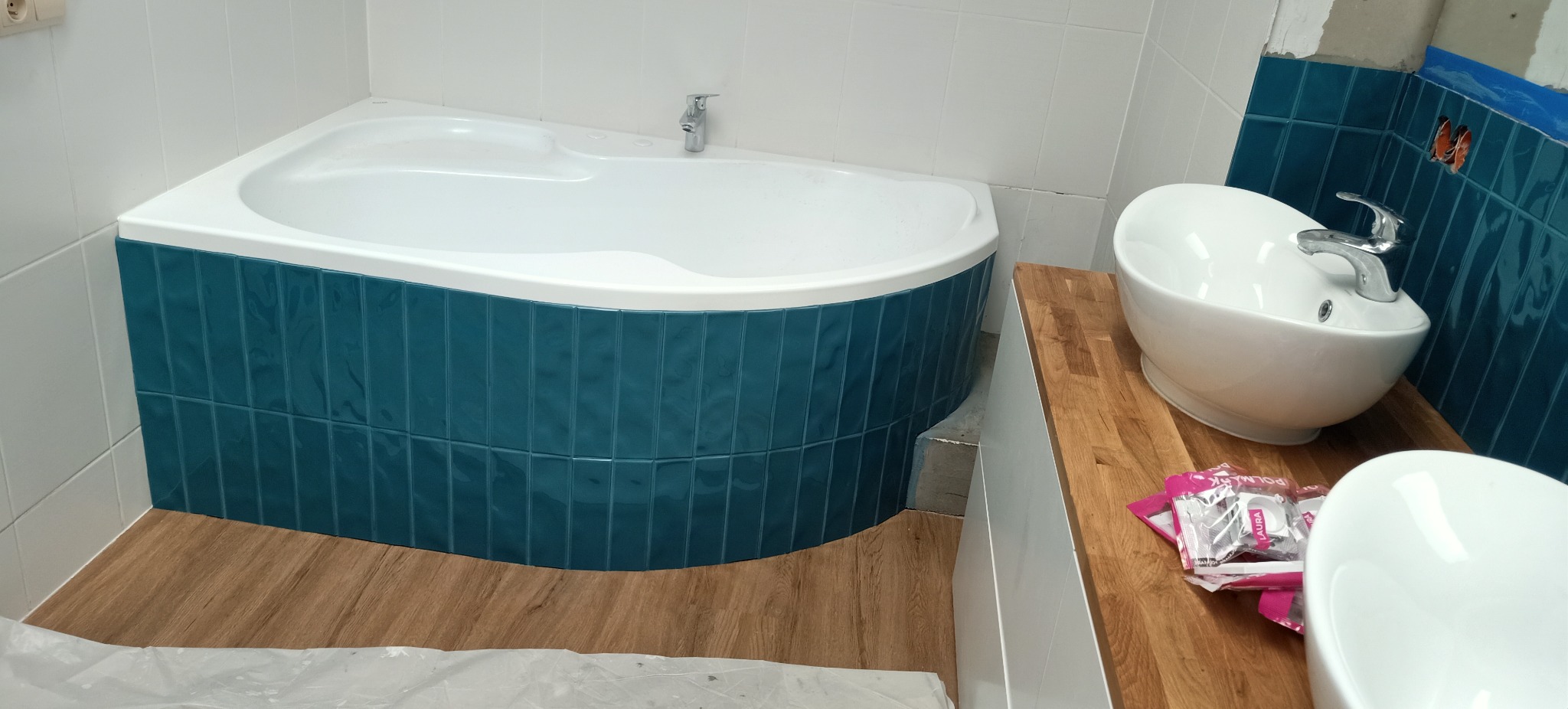 Narożna wanna obłożona turkusowymi, pionowo ułożonymi płytkami, umywalka nablatowa na drewnianym blacie, częściowo wykończona ściana z turkusową glazurą w trakcie remontu łazienki.