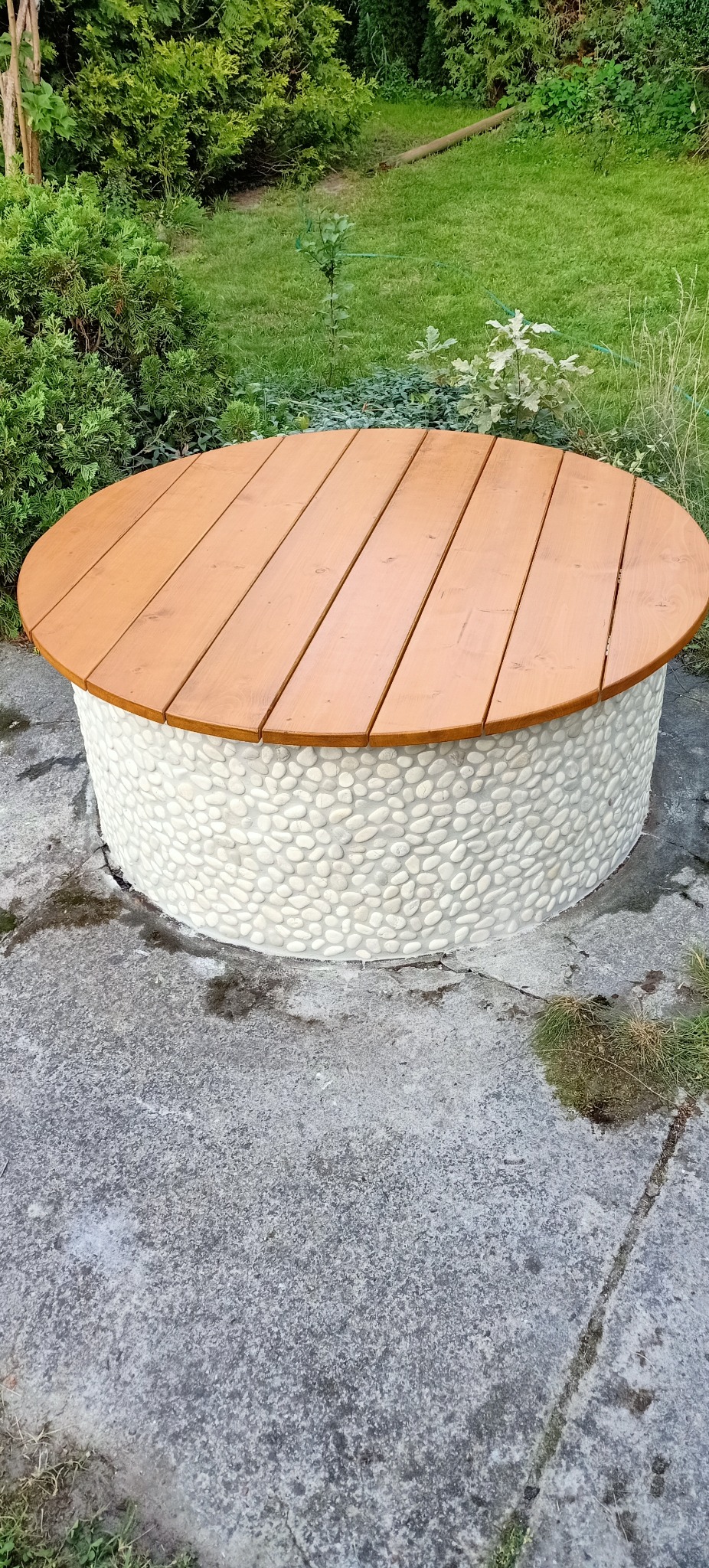 Okrągły stolik z drewnianym blatem i podstawą wykończoną białymi kamykami na betonowym podłożu, otoczony zielenią.