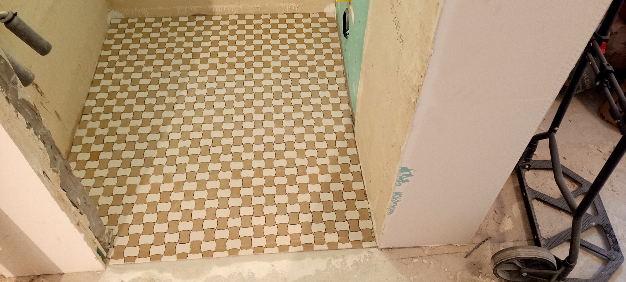Wykończona podłoga w łazience z beżowo-białą mozaiką ułożoną w geometryczny wzór; widoczne surowe ściany w trakcie prac wykończeniowych i wózek transportowy.