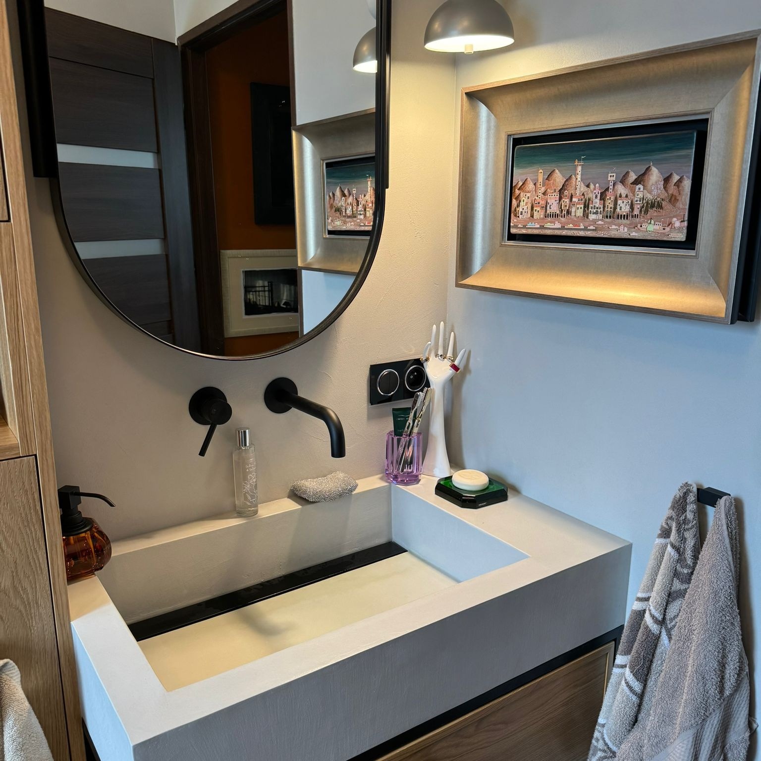 Nowoczesna łazienka z prostokątną umywalką z betonu architektonicznego, czarną armaturą i okrągłym lustrem, ozdobiona obrazem w złotej ramie i akcesoriami w odcieniach fioletu i bieli.