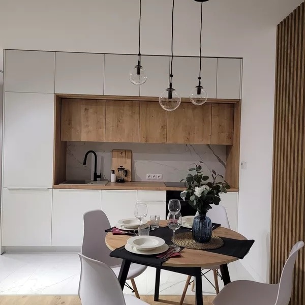 Nowoczesna kuchnia z białymi szafkami, drewnianymi akcentami i okrągłym stołem z nakryciem, oświetlona trzema wiszącymi lampami z przezroczystymi kulami.