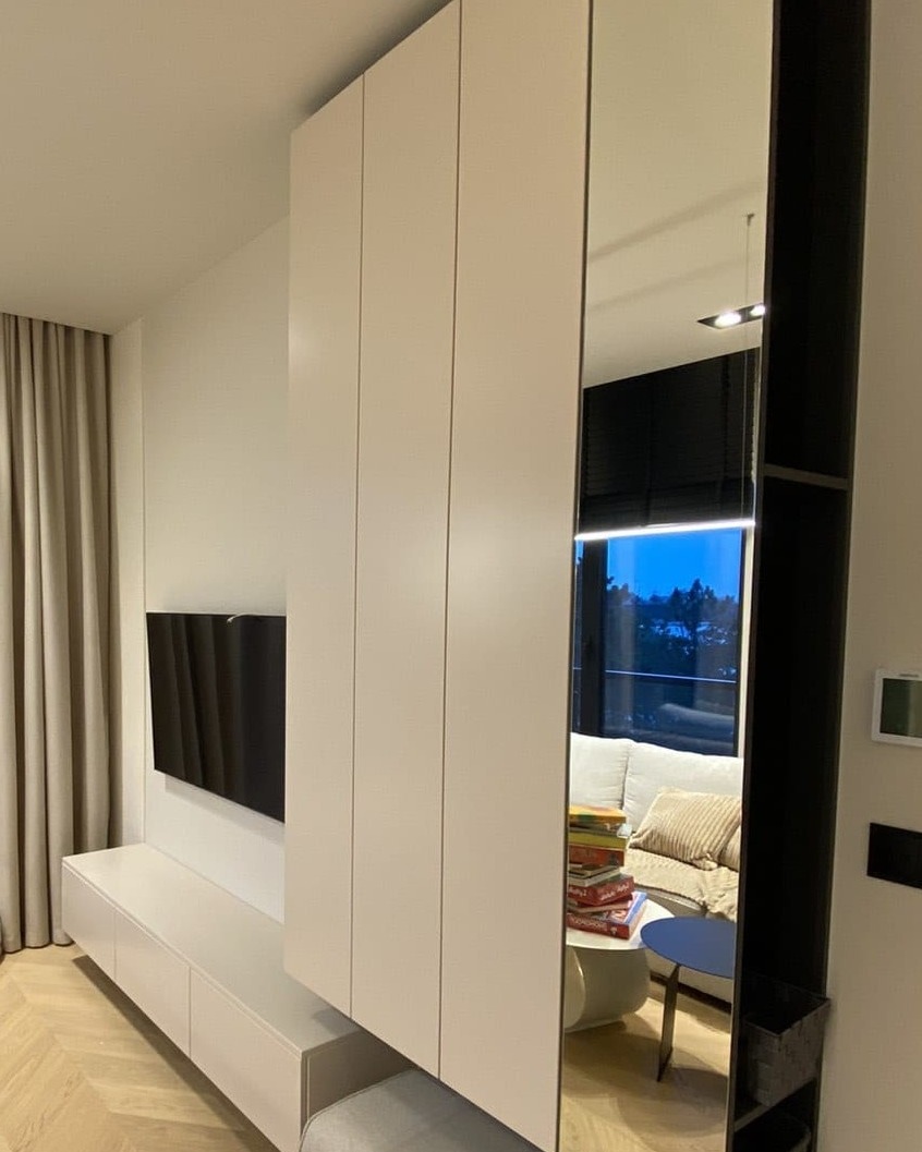 Nowoczesny salon z białymi meblami na wymiar, wiszącym telewizorem i lustrem w czarnej ramie odbijającym wnętrze z kanapą i stosem książek na stoliku.