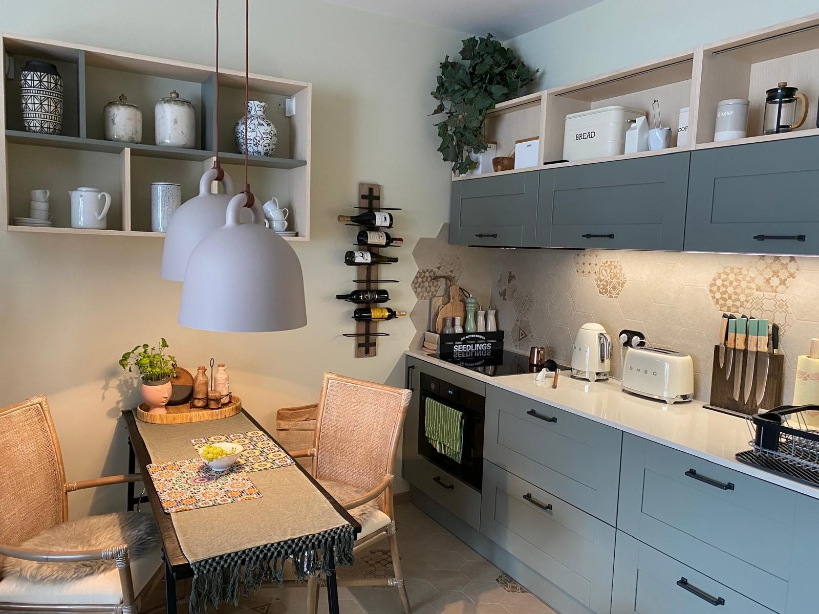 Jasna kuchnia z szarymi szafkami, białym blatem, heksagonalnymi płytkami na ścianie, drewnianym regałem na wino, wiszącymi lampami i stołem z nakryciem w stylu vintage.