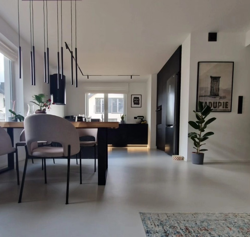 Jasne wnętrze z otwartą kuchnią, drewnianym stołem i krzesłami w odcieniach szarości, minimalistyczne oświetlenie wiszące, czarne meble kuchenne z podświetleniem LED, plakat na ścianie i roślina...