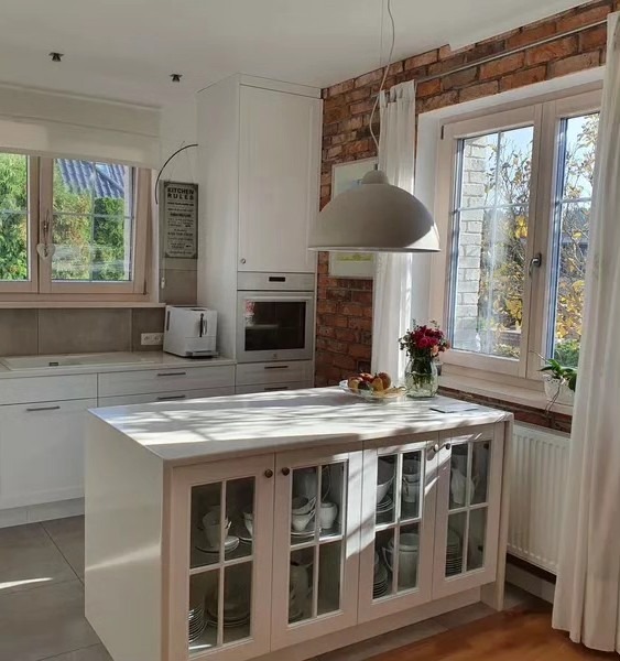 Jasna kuchnia z wyspą z przeszklonymi frontami szafek, ceglana ściana nad blatem, owocami w misie i bukietem kwiatów na wyspie, oraz wiszącą lampą w kształcie kopuły.