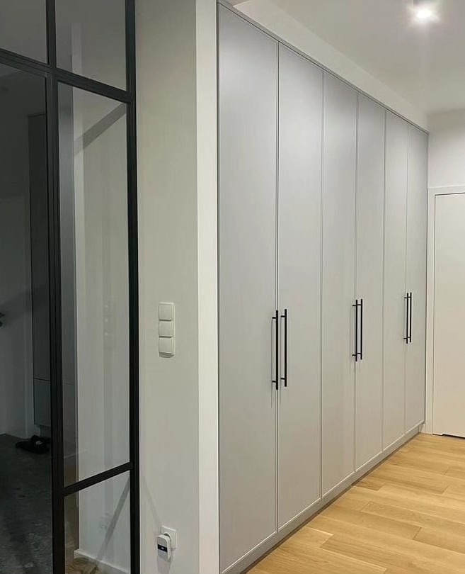 Długa, szara szafa wnękowa z czarnymi, pionowymi uchwytami, przylegająca do ściany w korytarzu z parkietem i czarną, przeszkloną ścianką działową.