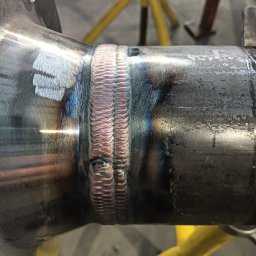 SpawMat Kamil Matysik - Metalowy element konstrukcyjny po procesie spawania. Widoczne ślady spawu z charakterystyczną strukturą. W tle fragment żółtego stojaka warsztatowego.