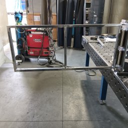 SpawMat Kamil Matysik - Metalowa konstrukcja w warsztacie, widoczna spawarka Lincoln Electric, stół spawalniczy z narzędziami. Rama w trakcie tworzenia, surowe wykończenie.