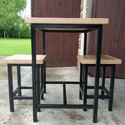 SpawMat Kamil Matysik - Metalowy stół barowy z dwoma stołkami, czarne profile, drewniany blat i siedziska, na zewnątrz, brązowe drzwi w tle.