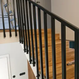 Nowoczesna, czarna poręcz schodowa z pionowymi szczeblami na drewnianych schodach. Widoczny fragment drabiny w tle. Minimalistyczny design.