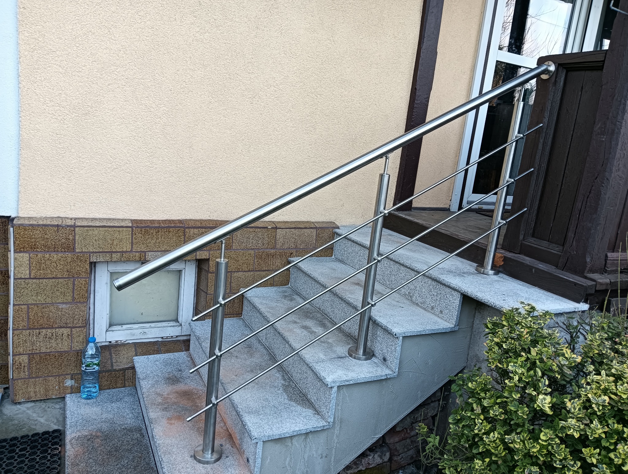 Zewnętrzne schody z szarego kamienia z nowoczesną, minimalistyczną poręczą ze stali nierdzewnej. Butelka wody przy oknie piwnicznym.