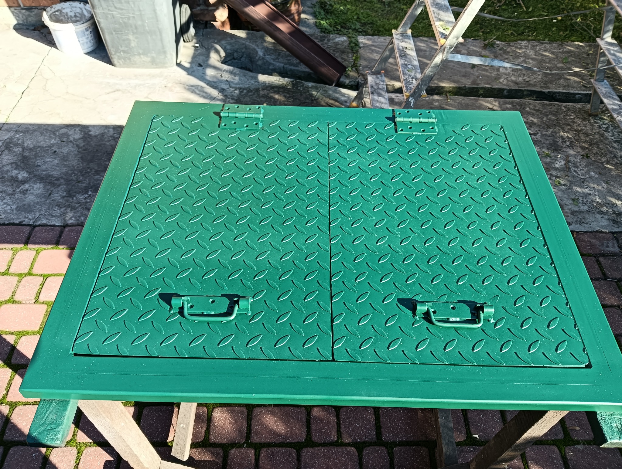 Zielona, metalowa klapa z uchwytami na stole, z fakturą łezki. Widoczne zawiasy. Pomalowana świeżą farbą. W tle kostka brukowa i drabina.