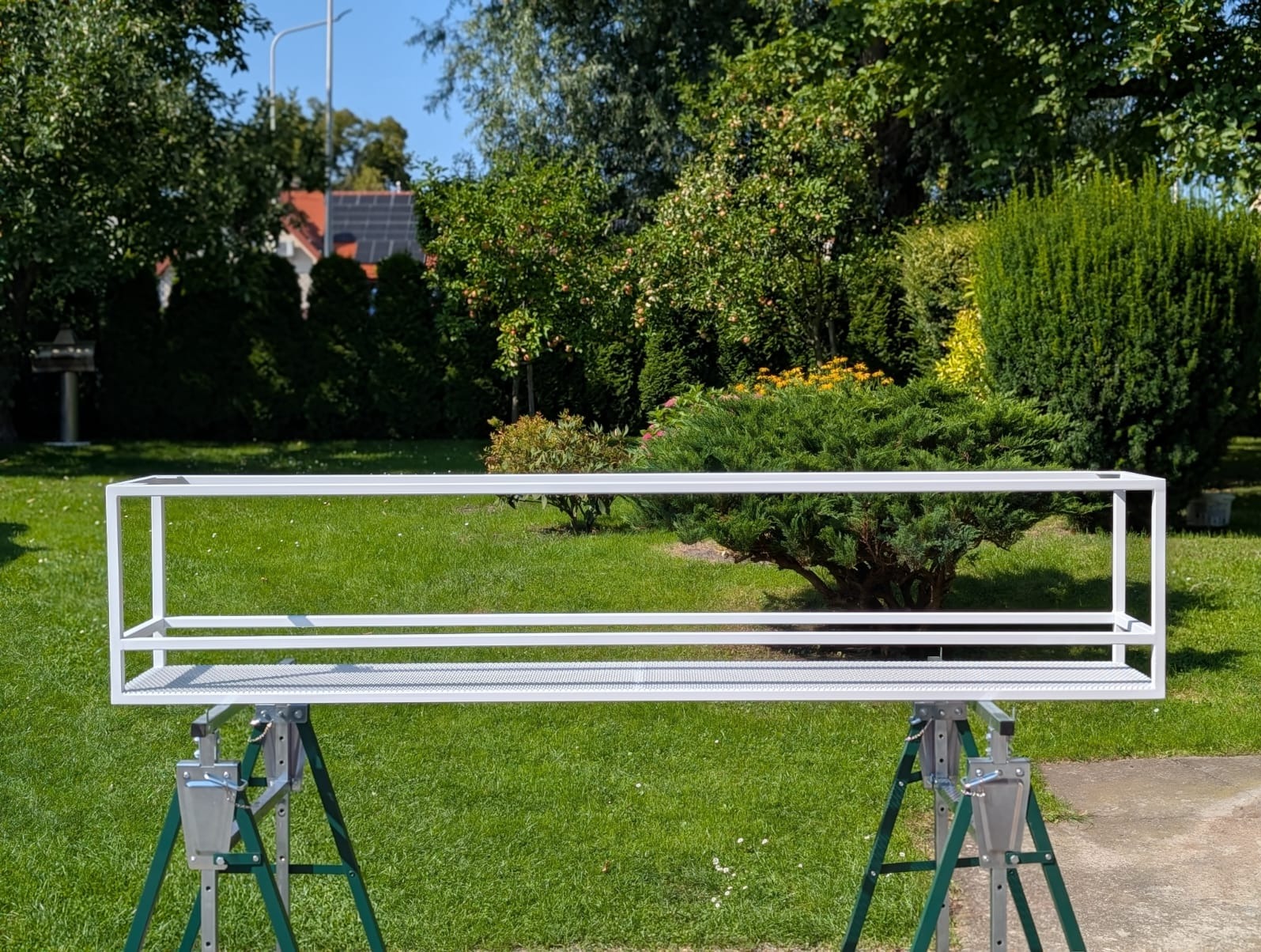 Biała, metalowa konstrukcja na zielonych kozłach, prawdopodobnie element zabudowy balkonu, na tle bujnej zieleni ogrodu w słoneczny dzień.