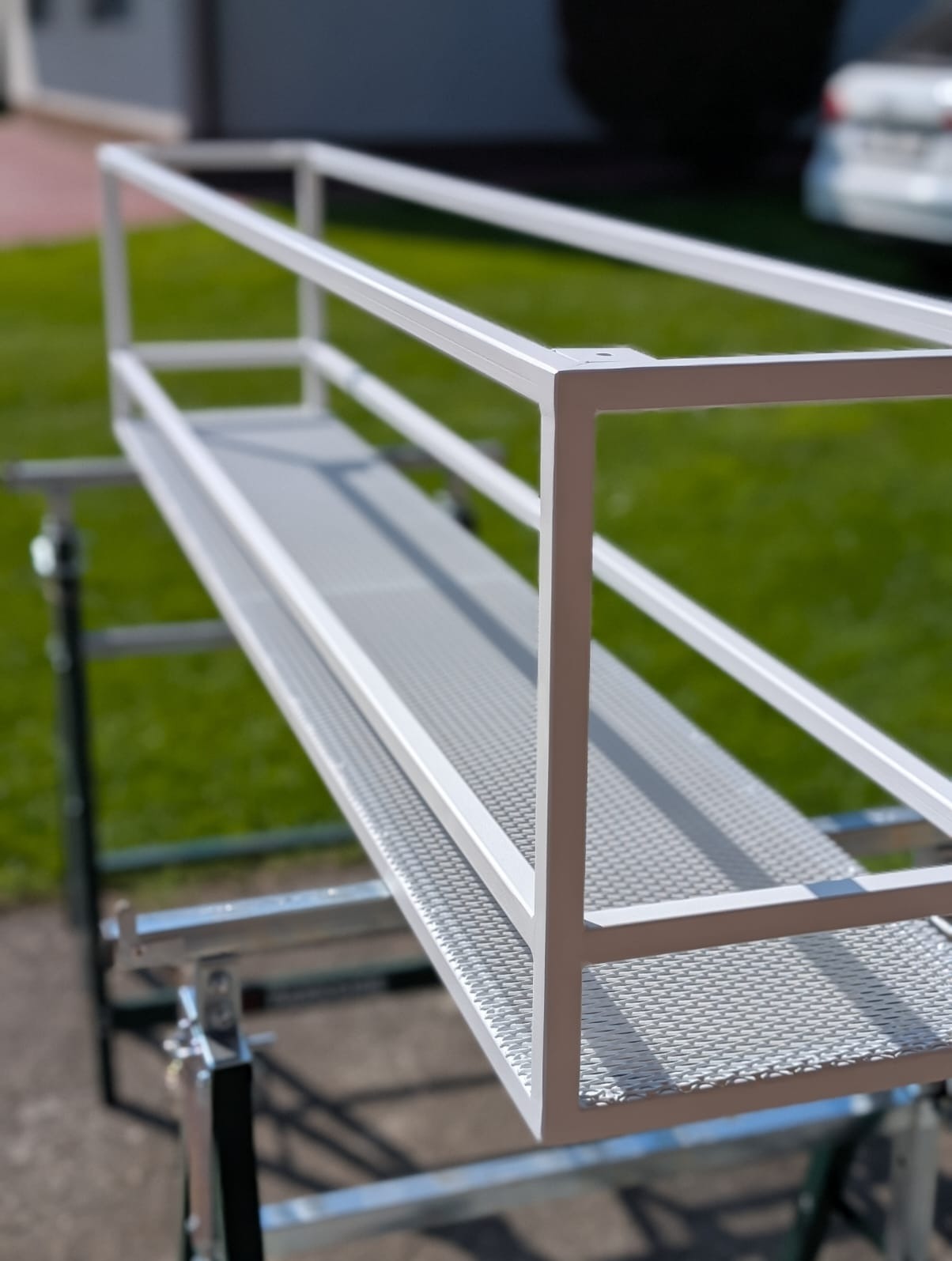 Biała, metalowa konstrukcja przypominająca szkielet balkonu lub pergoli, ustawiona na rusztowaniu na tle trawnika i domu. Widoczne detale spawów i siatki.