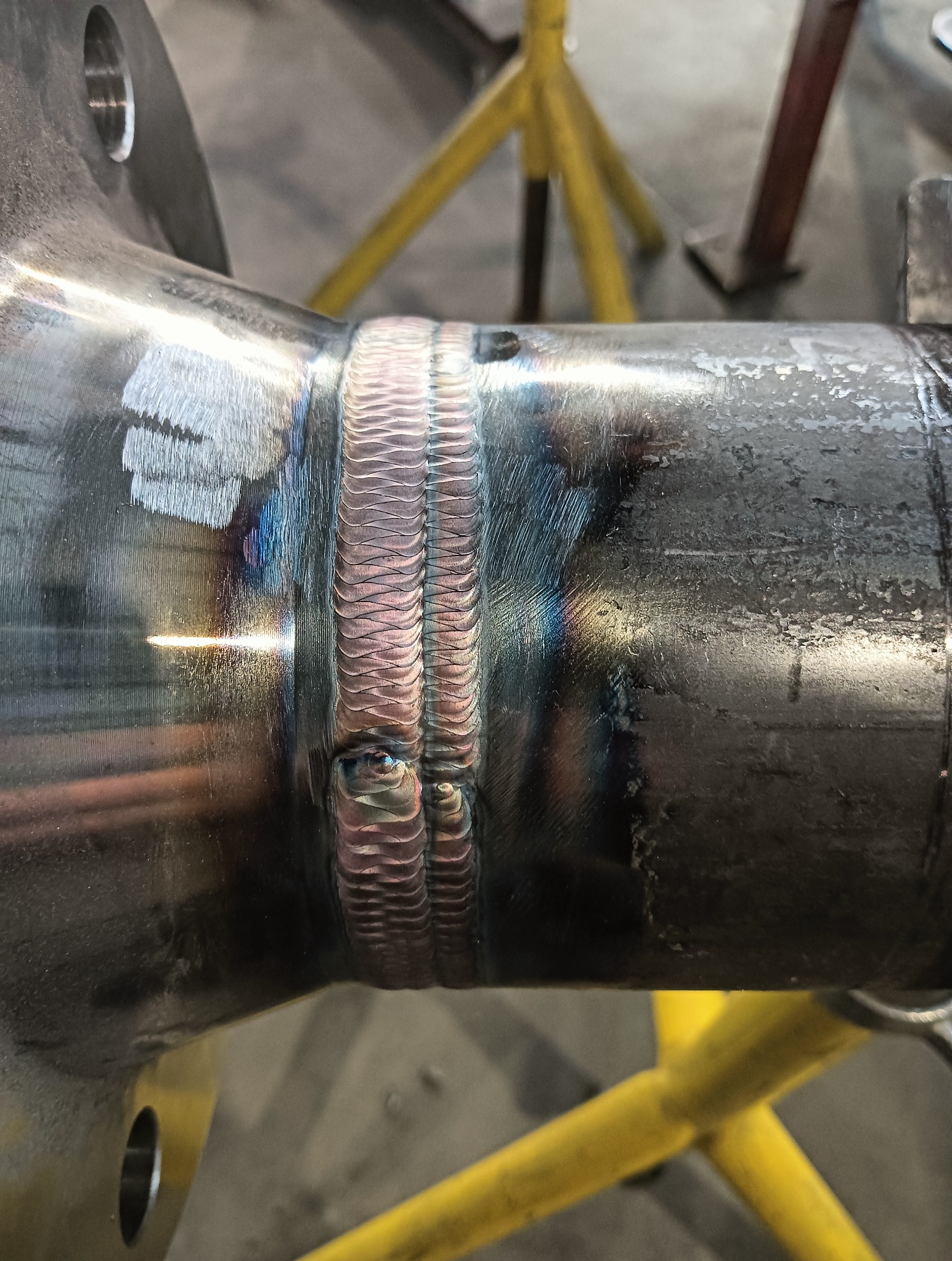 Metalowy element konstrukcyjny po procesie spawania. Widoczne ślady spawu z charakterystyczną strukturą. W tle fragment żółtego stojaka warsztatowego.