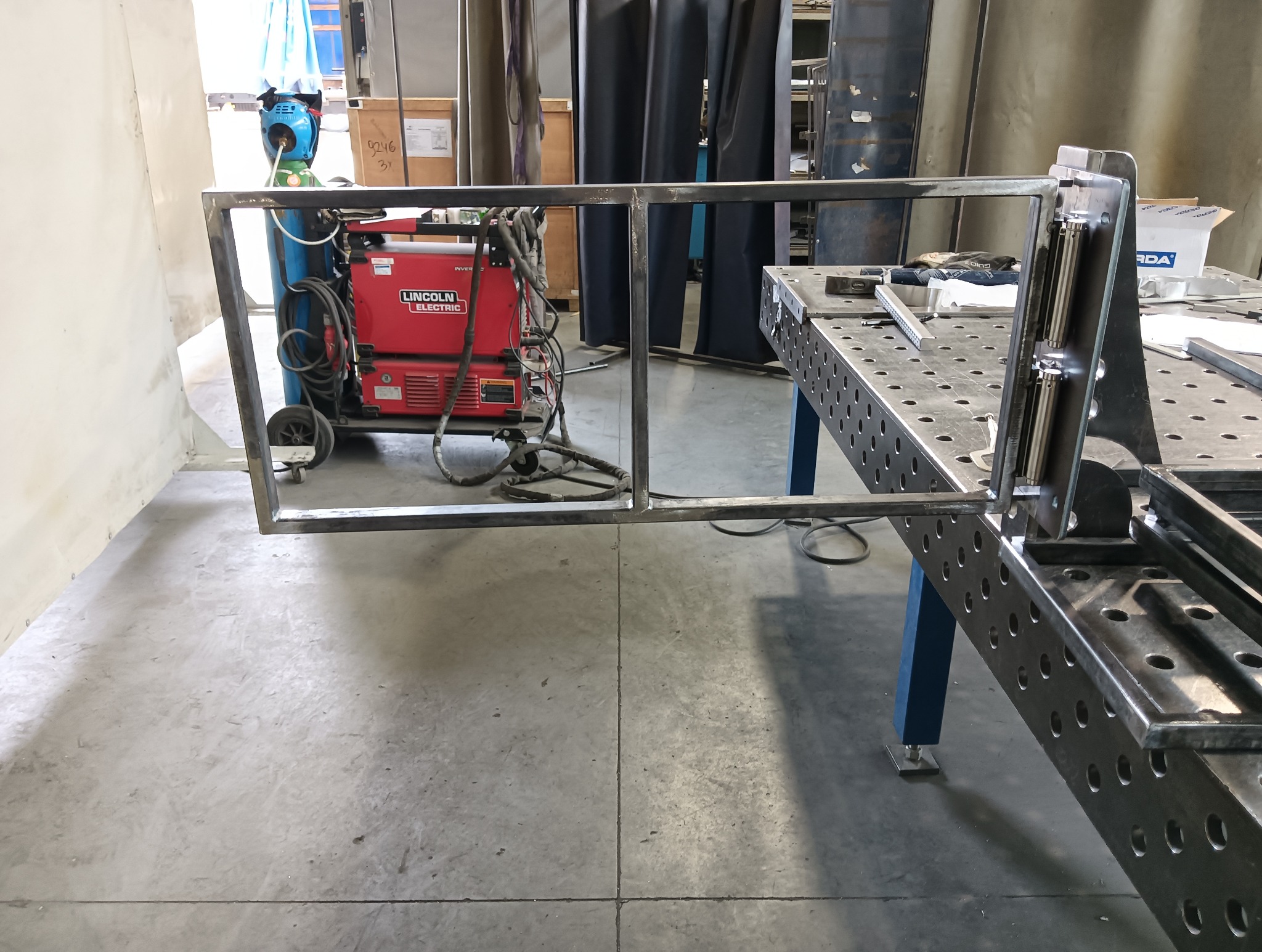 Metalowa konstrukcja w warsztacie, widoczna spawarka Lincoln Electric, stół spawalniczy z narzędziami. Rama w trakcie tworzenia, surowe wykończenie.