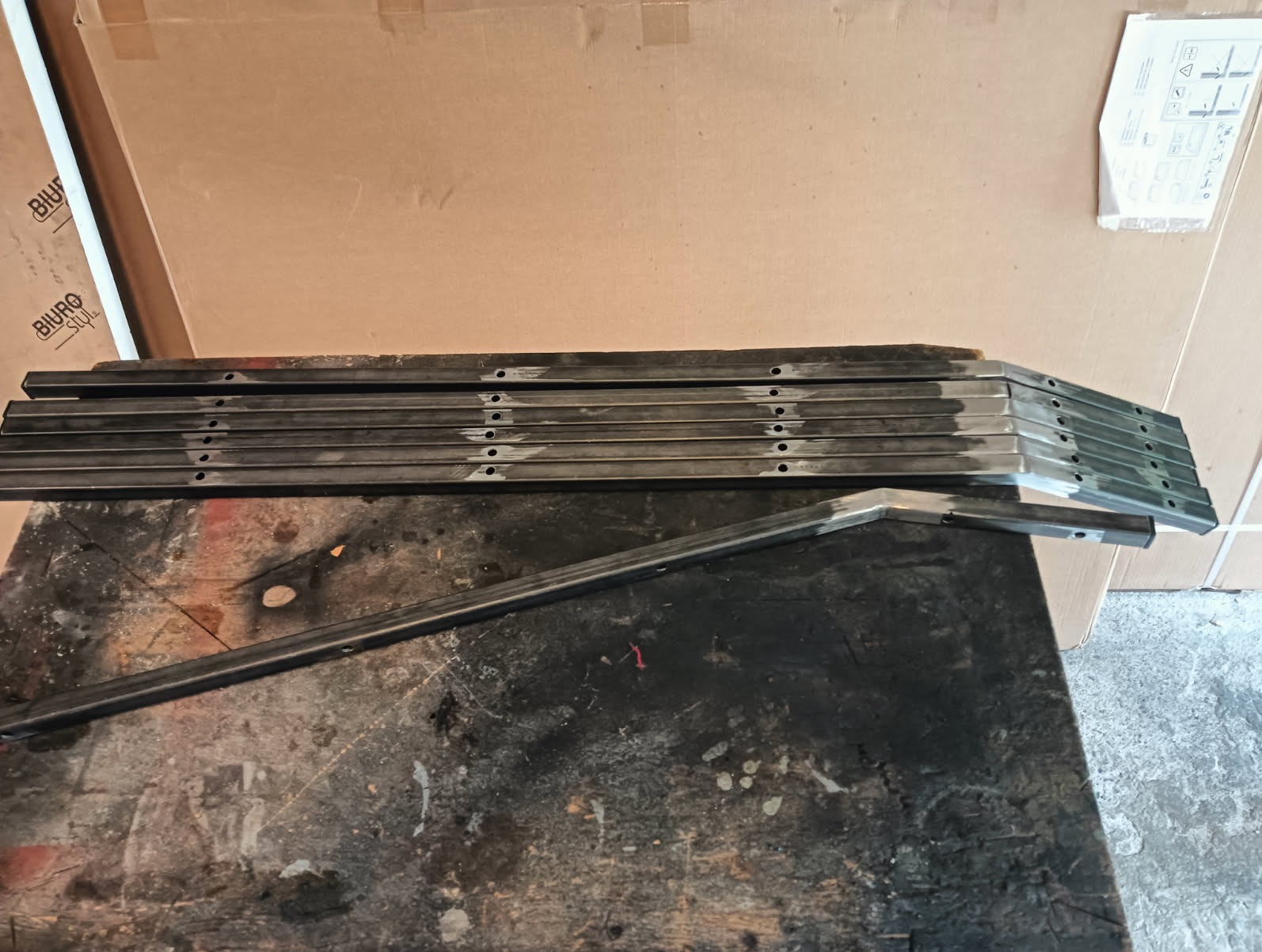 Stos metalowych profili z otworami, prawdopodobnie elementy konstrukcyjne, leżące na warsztatowym stole. Widoczne ślady obróbki.
