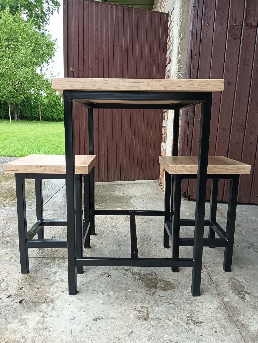 Metalowy stół barowy z dwoma stołkami, czarne profile, drewniany blat i siedziska, na zewnątrz, brązowe drzwi w tle.