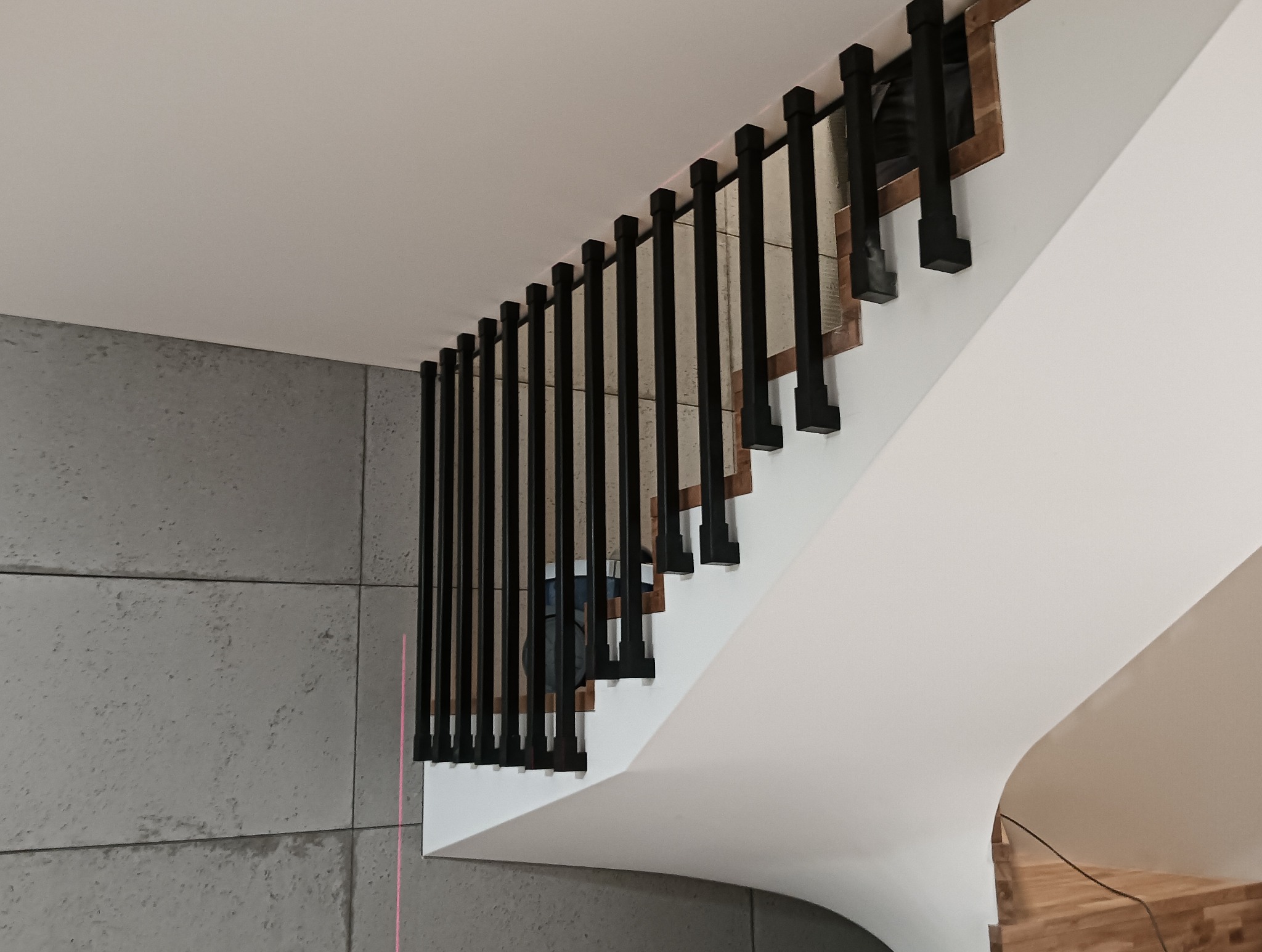 Nowoczesne, czarne poręcze schodowe z kwadratowymi elementami, zamontowane na białych stopniach, z szarą ścianą w tle. Minimalistyczny design.