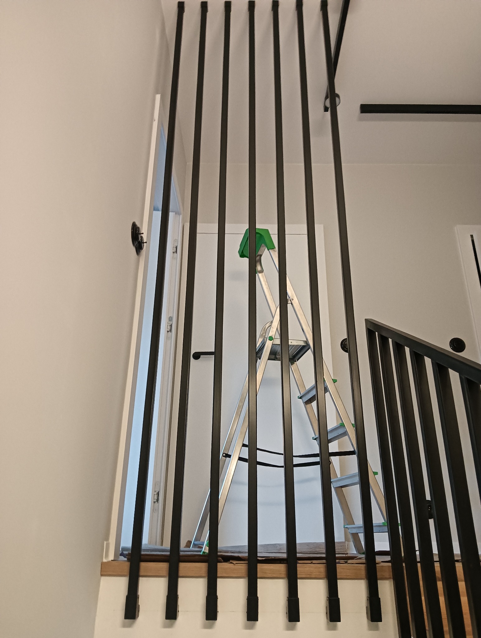 Nowoczesna, pionowa balustrada schodowa z czarnych, metalowych prętów. W tle biała ściana i drabina. Minimalistyczny design z akcentem na geometrię.